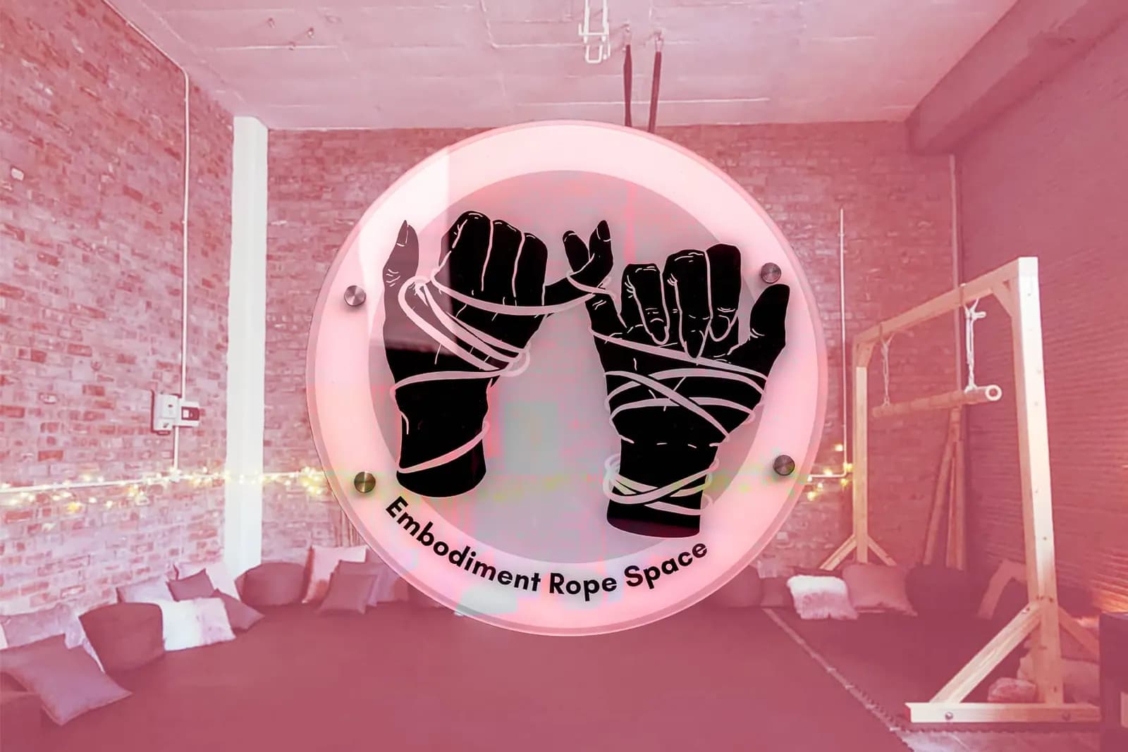 Sign with bound hands labeled "Embodiment Rope Space," cozy room with cushions, brick wall, and wood