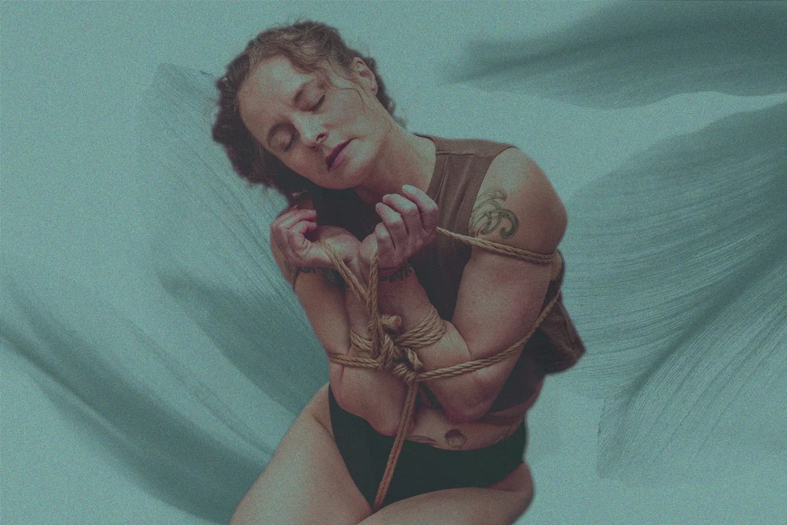 A person with eyes closed kneels with their arms and hands bound in front of their chest with jute rope, against a soft teal background.